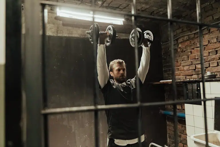 Man lifting dumbbells in a rustic gym with metal bars, showcasing strength training.